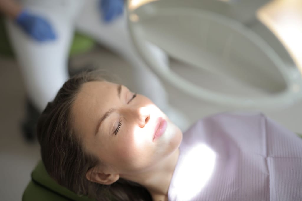 Calm patient reclining in a treatment chair with eyes closed, soft light; clinician only as out-of-focus gloved hands.
