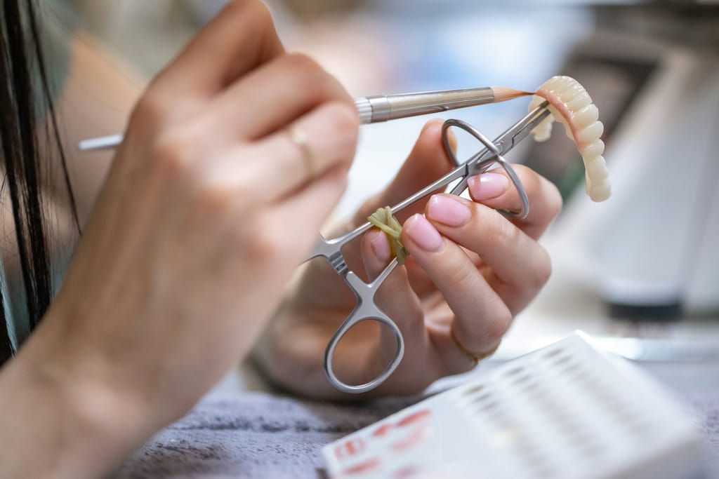 Hands with tweezers/clamp and a modelling instrument working on a ceramic bridge at a bench.